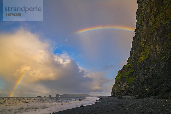 Die als Reynisdrangar bekannten Meeresstapel mit Wellen bei Sonnenaufgang und Tausenden von Vögeln  die in der Luft fliegen  mit einem Regenbogen  Südküste; Island'