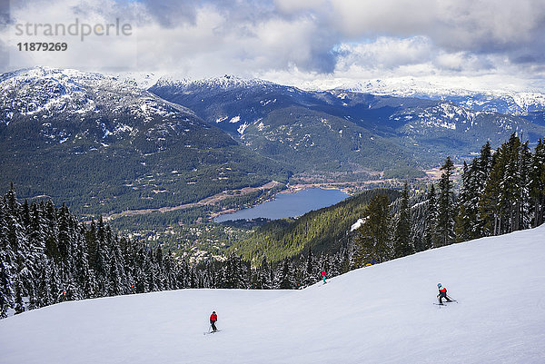 Skifahrer und Snowboarder  die einen Skihügel hinunterfahren  mit herrlichem Blick auf einen See und die kanadischen Rocky Mountains; Whistler  British Columbia  Kanada'.
