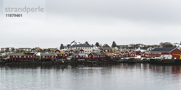 Rote Gebäude entlang der Wasserkante bei bewölktem Himmel; Lofoten  Nordland  Norwegen'.
