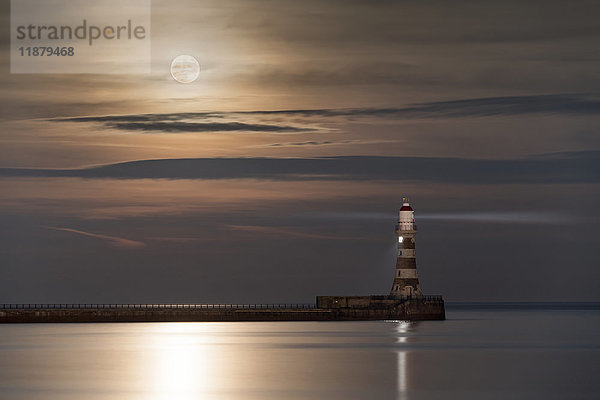 Roker Lighthouse leuchtet am Ende eines Piers unter einem hellen Vollmond  der sich auf dem ruhigen Wasser spiegelt; Sunderland  Tyne and Wear  England