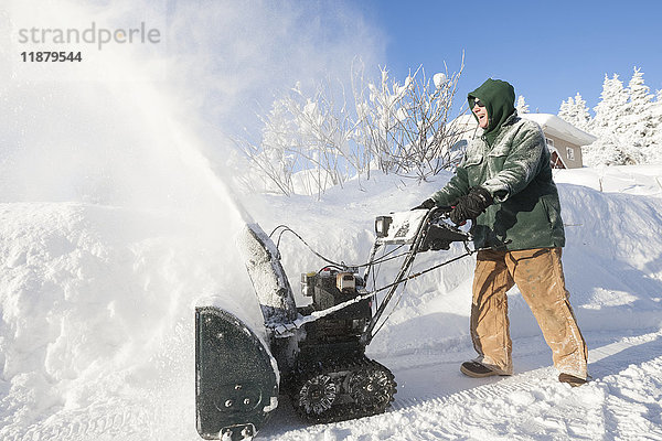 Ein Mann benutzt fröhlich seine Schneefräse vor seinem Haus; Alaska  Vereinigte Staaten von Amerika'.