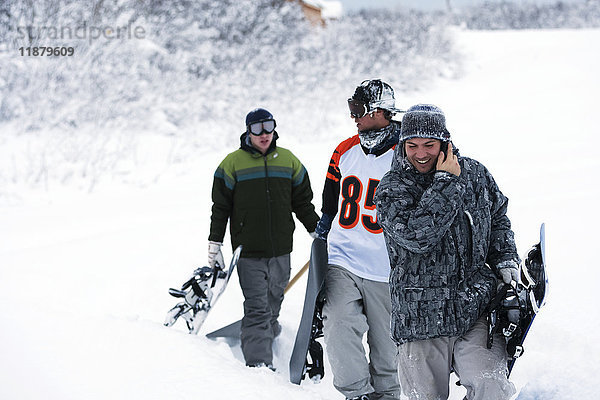 Drei Snowboarder  die mit ihren Snowboards eine Piste im Tiefschnee hinaufgehen  ein Snowboarder spricht mit einem Handy; Alaska  Vereinigte Staaten von Amerika