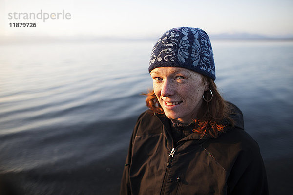 Porträt einer Frau mit roten Haaren  die eine Strickmütze trägt und mit Wasser im Hintergrund steht; Alaska  Vereinigte Staaten von Amerika