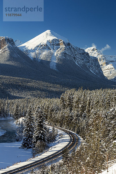 Schneebedeckte Bergkette mit vereisten und schneebedeckten immergrünen Bäumen entlang eines Flusses mit Eisenbahnschienen und blauem Himmel; Banff  Alberta  Kanada'.