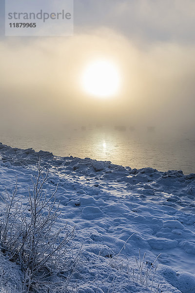 Die Sonne scheint durch den tief liegenden Nebel und taucht den Turnagain Arm und den Seward Highway in ein warmes Licht  während im Vordergrund das Meereis den Ozean bedeckt  Süd-Zentral-Alaska; Anchorage  Alaska  Vereinigte Staaten von Amerika'.