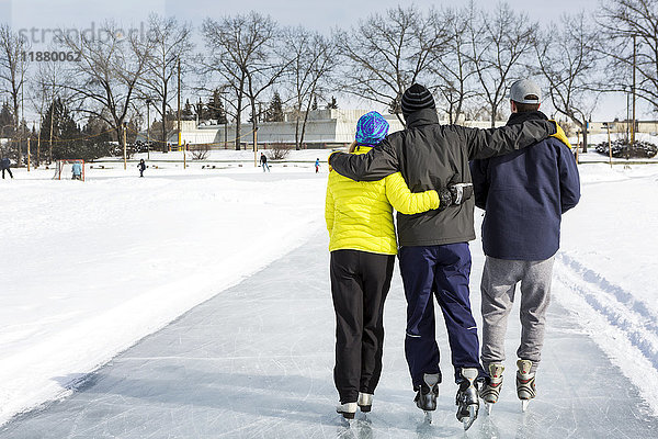 Zwei Männer und eine Frau laufen Arm in Arm auf frisch präpariertem Eis auf einem Teich mit einem Gemeindezentrum im Hintergrund; Calgary  Alberta  Kanada'.