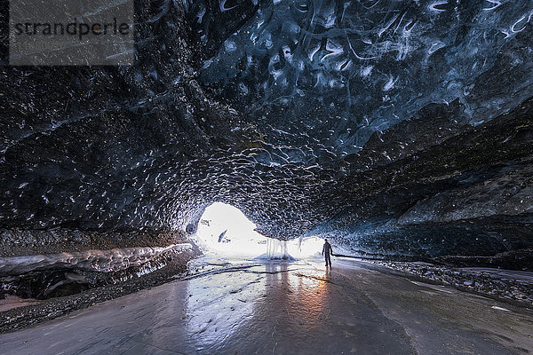 Ein Mann steht in der Nähe des Eingangs zu einem Tunnel unter dem Eis des Canwell-Gletschers. Die Farben des Sonnenuntergangs spiegeln sich auf dem Eis  mitten im Winter; Alaska  Vereinigte Staaten von Amerika