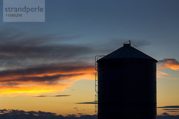 Silhouette eines großen Getreidesilos aus Metall mit bunten Wolken bei Sonnenuntergang und blauem Himmel; Alberta  Kanada'.