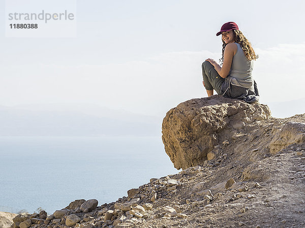 Eine junge Frau sitzt auf einem Felsen  blickt zurück und posiert für die Kamera mit einem wolkenverhangenen Tal unter ihr  Naturreservat Ein Gedi  Bezirk Totes Meer; Region Süd  Israel '