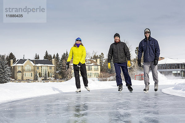 Zwei Männer und eine Frau laufen Schlittschuh auf frisch präpariertem Eis auf einem Teich mit Häusern im Hintergrund; Calgary  Alberta  Kanada'.