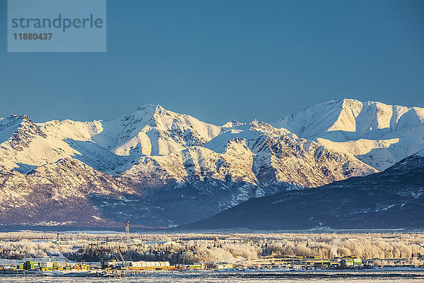 Blick auf die Innenstadt von Anchorage im Winter und die schneebedeckten Chugach Mountains von Point MacKenzie aus gesehen  der Himmel im Hintergrund ist klar und hell  das Meereis bedeckt das Cook Inlet im Vordergrund  Süd-Zentral-Alaska; Anchorage  Alaska  Vereinigte Staaten von Amerika'.