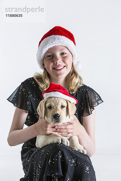 Ein lächelndes junges Mädchen mit Weihnachtsmannmütze umarmt einen Labradorwelpen  der ebenfalls eine Weihnachtsmannmütze trägt  vor einem weißen Hintergrund; Anchorage  Alaska  Vereinigte Staaten von Amerika'.