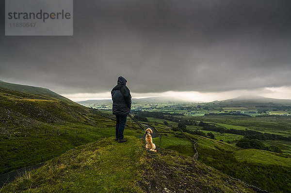 Ein Mann steht mit seinem Hund auf einem grasbewachsenen Hügel und blickt auf die üppige  grüne Landschaft unter einem stürmischen Himmel; North Yorkshire  England'.
