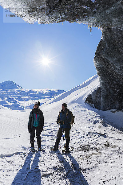Ein Paar  das mit Schneeschuhen unterwegs ist und an einem sonnigen Wintertag unter dem Eingang einer Eishöhle am Canwell-Gletscher in der Alaska Range steht; Alaska  Vereinigte Staaten von Amerika'.