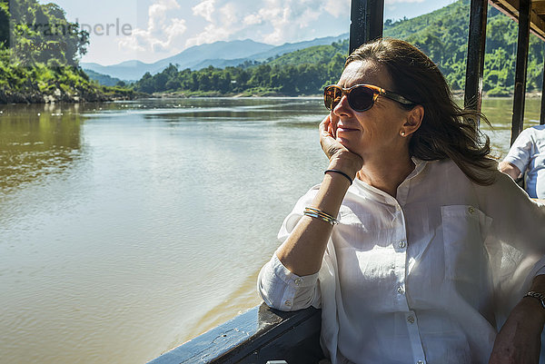Eine Frau mit Sonnenbrille sitzt in einem Ausflugsboot und schaut aus dem Fenster  während sie den Mekong hinunterfährt; Provinz Luang Prabang  Laos'.