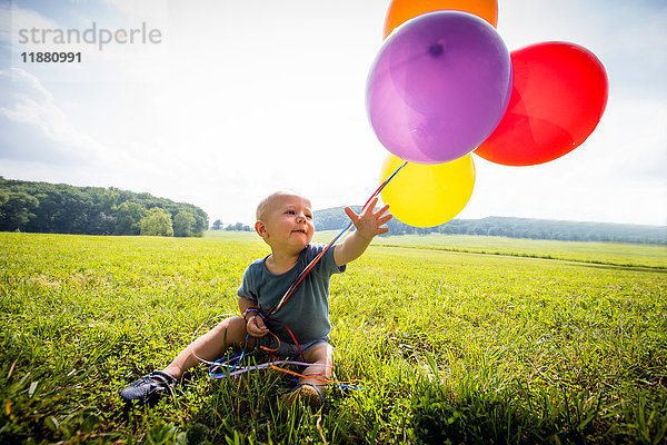 Kleiner Junge sitzt mit einem Haufen bunter Luftballons auf einem ländlichen Feld