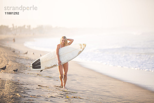 Porträt einer jungen Surferin  die ein Surfbrett am Strand entlang trägt  Santa Monica  Kalifornien  USA