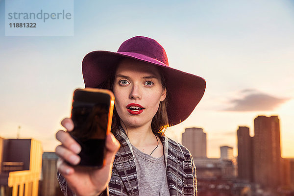 Porträt einer jungen Frau  die sich auf einer Dachparty bei Sonnenuntergang in London  UK  mit einem Smartphone selbstständig macht