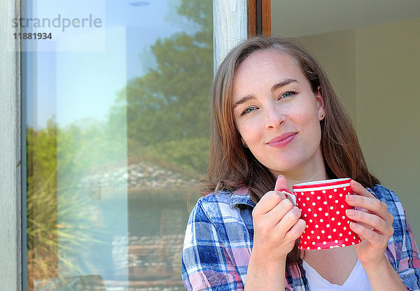 Porträt einer jungen Frau  die eine Kaffeetasse an der Terrassentür hält