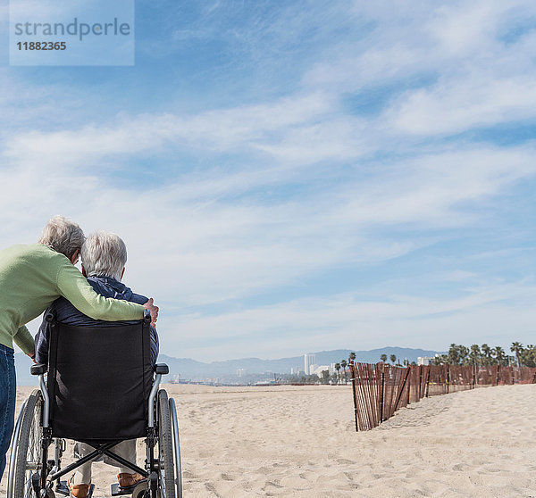 Rückansicht eines älteren Mannes im Rollstuhl mit Frau  der vom Strand aus auf den Strand schaut  Santa Monica  Kalifornien  USA