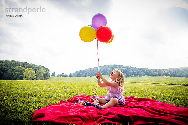Mädchen sitzt auf roter Decke in ländlichem Feld und schaut auf einen Haufen bunter Luftballons