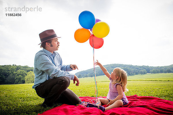 Mädchen sitzt mit dem Vater zusammen und zeigt auf einen Haufen bunter Luftballons auf einem ländlichen Feld