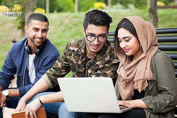 Drei Freunde  die auf einer Parkbank sitzen und auf einen Laptop schauen