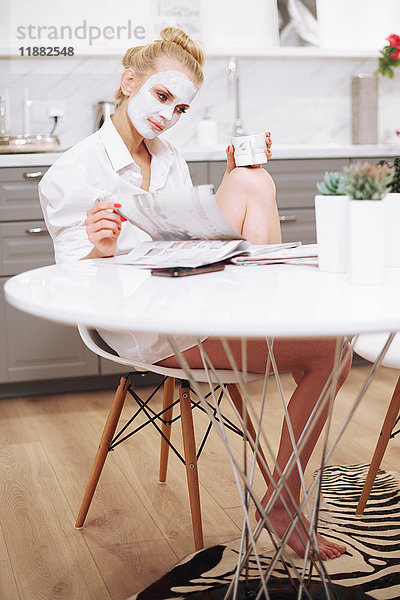 Frau mit Gesichtsmaske sitzt am Esstisch und liest Zeitschrift