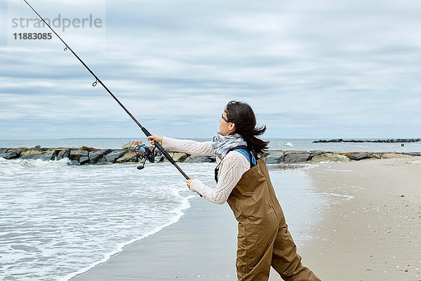 Junge Frau in Watvögeln beim Auswerfen der Meeresangel vom Strand