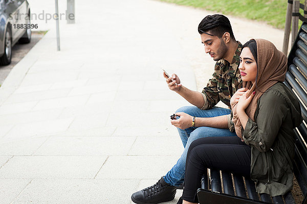 Junges Paar sitzt auf Parkbank  junger Mann schaut auf Smartphone
