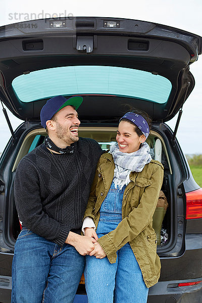 Junges Paar sitzt im offenen Kofferraum eines Autos  hält Händchen und lacht