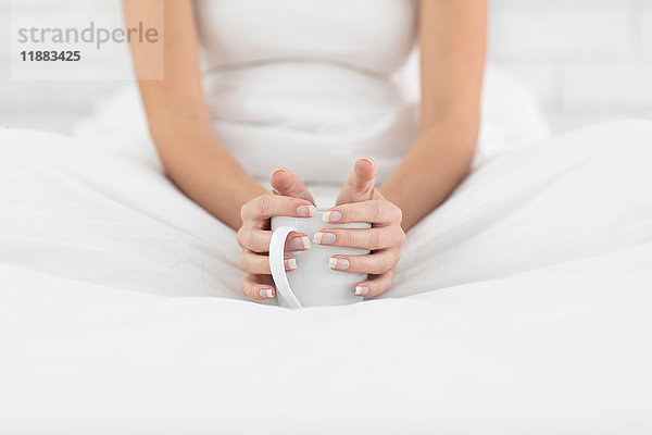 Junge Frau sitzt im Bett  hält eine Tasse Kaffee in der Hand  Mittelteil