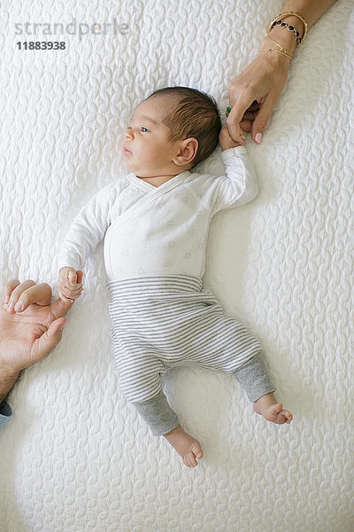 Auf dem Bett liegender Junge  Mutter und Vater halten die Hände des Babys  Blick von oben