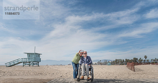 Älterer Mann im Rollstuhl mit Frau  die vom Strand aus auf ihn zeigt  Santa Monica  Kalifornien  USA