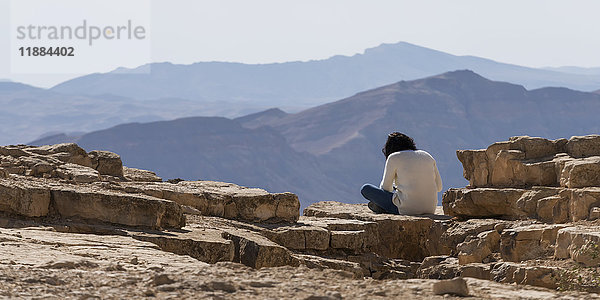 Eine Person sitzt auf einem Felsen mit Blick auf die zerklüftete Landschaft  Ramon-Naturreservat; Mitzpe Ramon  Südbezirk  Israel'.