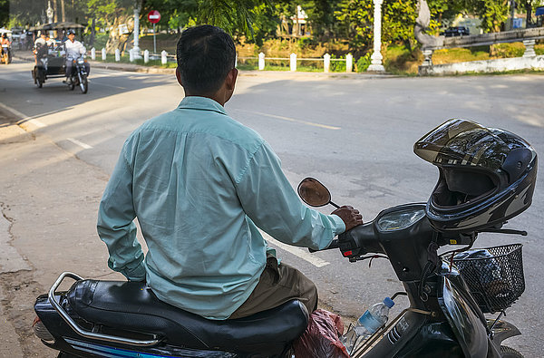 Ein Mann sitzt auf seinem Motorrad am Straßenrand  während sich eine Fahrradrikscha nähert; Krong Siem Reap  Provinz Siem Reap  Kambodscha'.