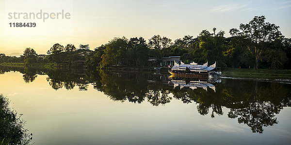 Spiegelungen in einem ruhigen Fluss bei Sonnenuntergang mit einem Ausflugsboot entlang des Ufers; Chiang Rai  Thailand'.
