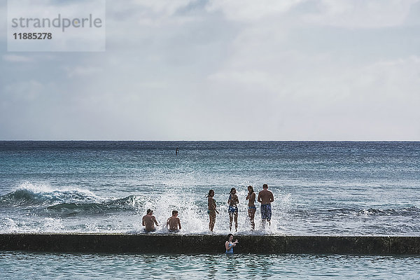 Touristen  die auf einer Mauer sitzen und stehen  während die Wellen mit Blick auf den Ozean hereinplatschen; Honolulu  Oahu  Hawaii  Vereinigte Staaten von Amerika'.