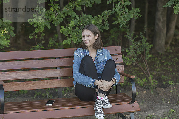 Lächelnde junge Frau sitzt auf einer Holzbank gegen Bäume im Park.
