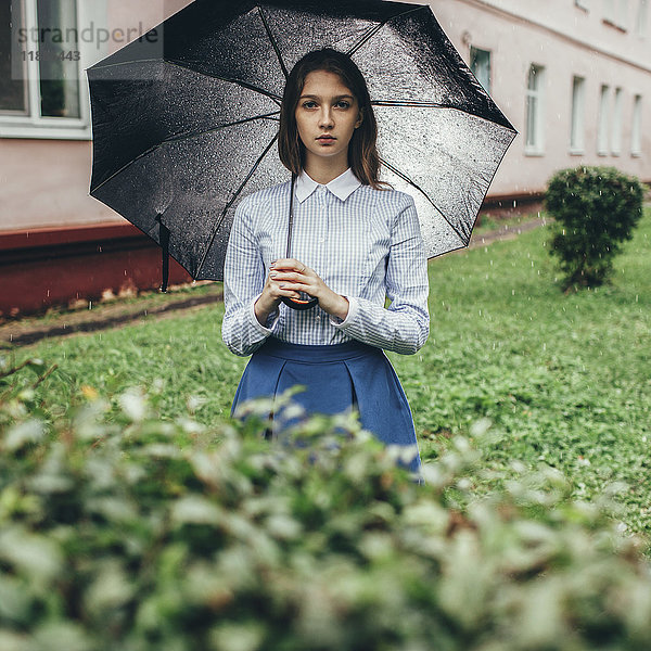 Porträt eines Teenagers  der bei Regen auf einer Wiese steht und einen Regenschirm hält.