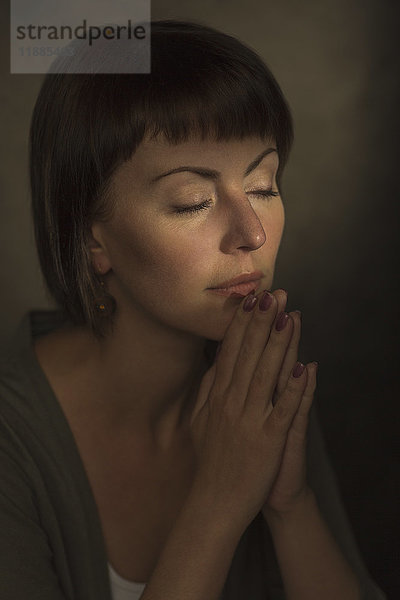 Mittlere erwachsene Frau mit geschlossenen Augen  die in Gebetsstellung vor grauem Hintergrund meditiert.
