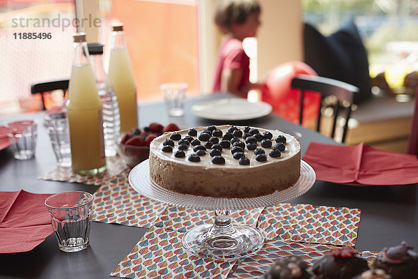 Torte auf Ständer über Platzmatten durch Flaschen am Tisch mit spielendem Mädchen im Hintergrund