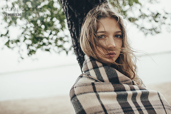 Porträt eines Teenagermädchens  eingehüllt in eine Decke gegen den See am windigen Tag.