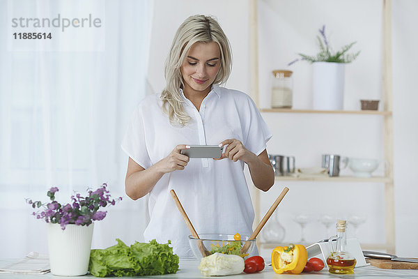 Lächelnde junge Frau fotografiert frischen Salat durch Smartphone in der Küche