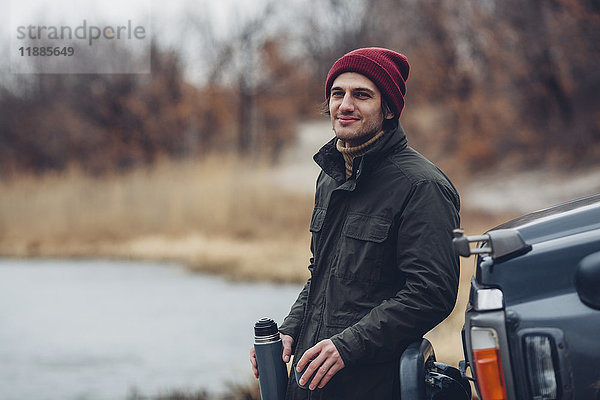 Lächelnder Mann hält isolierten Getränkebehälter  während er am Seeufer neben einem Geländewagen steht