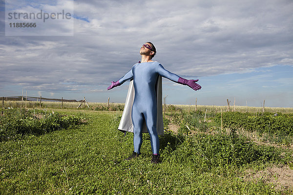 Mann in Superheldenkostüm stehend mit ausgestreckten Armen auf grasbewachsenem Feld