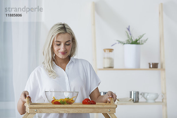 Schöne junge Frau hält Salatschüssel und Tomaten im Serviertablett in der Küche.