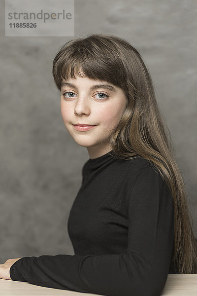 Porträt eines schönen lächelnden Mädchens mit langen Haaren am Tisch vor grauem Hintergrund