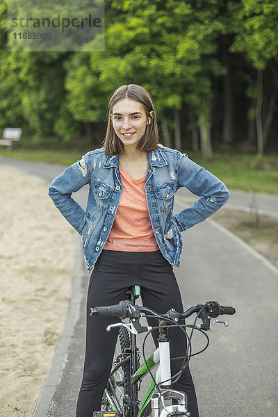 Lächelndes Porträt einer Frau mit Händen an der Hüfte  die auf dem Fahrrad auf der Straße im Park sitzt.