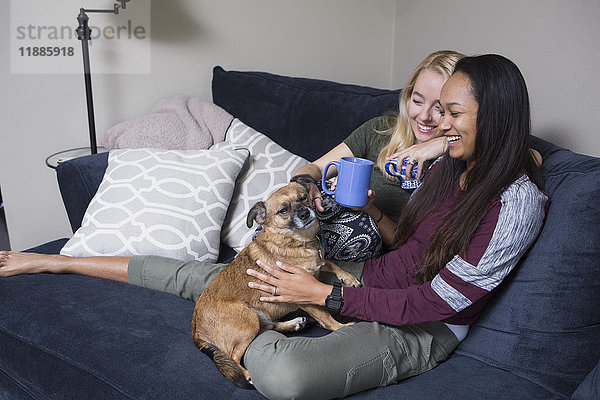 Fröhliche junge Frauen sitzen mit Kaffeetassen und Hund auf dem Sofa zu Hause.